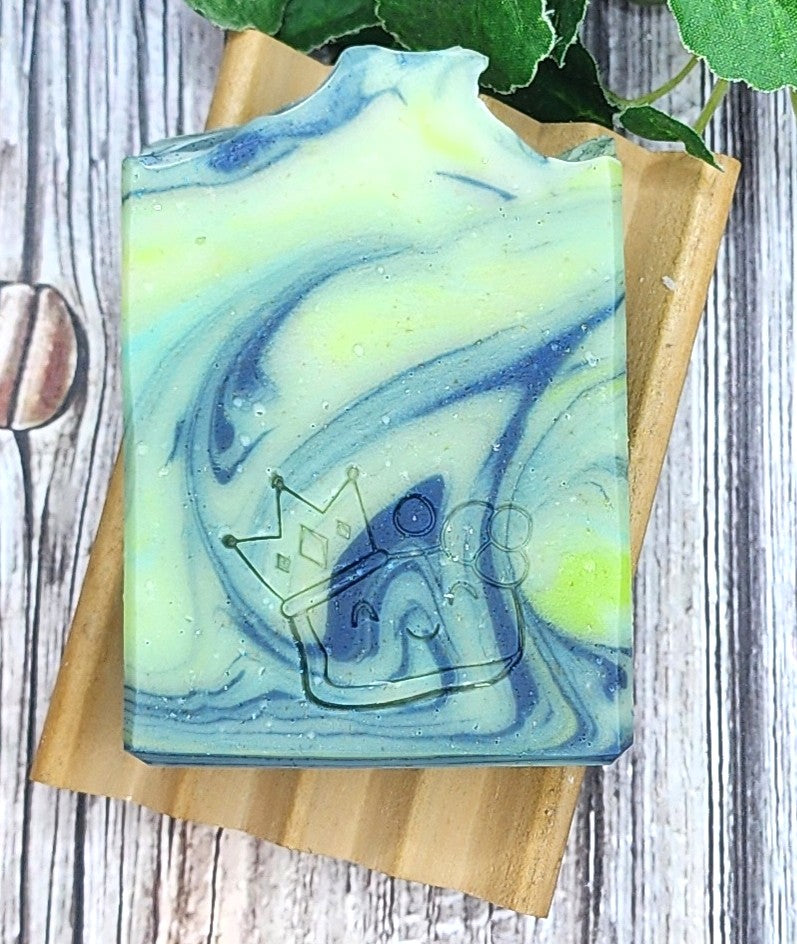 Marbled blue and green soap bar on a wooden soap dish with a plant in the background.