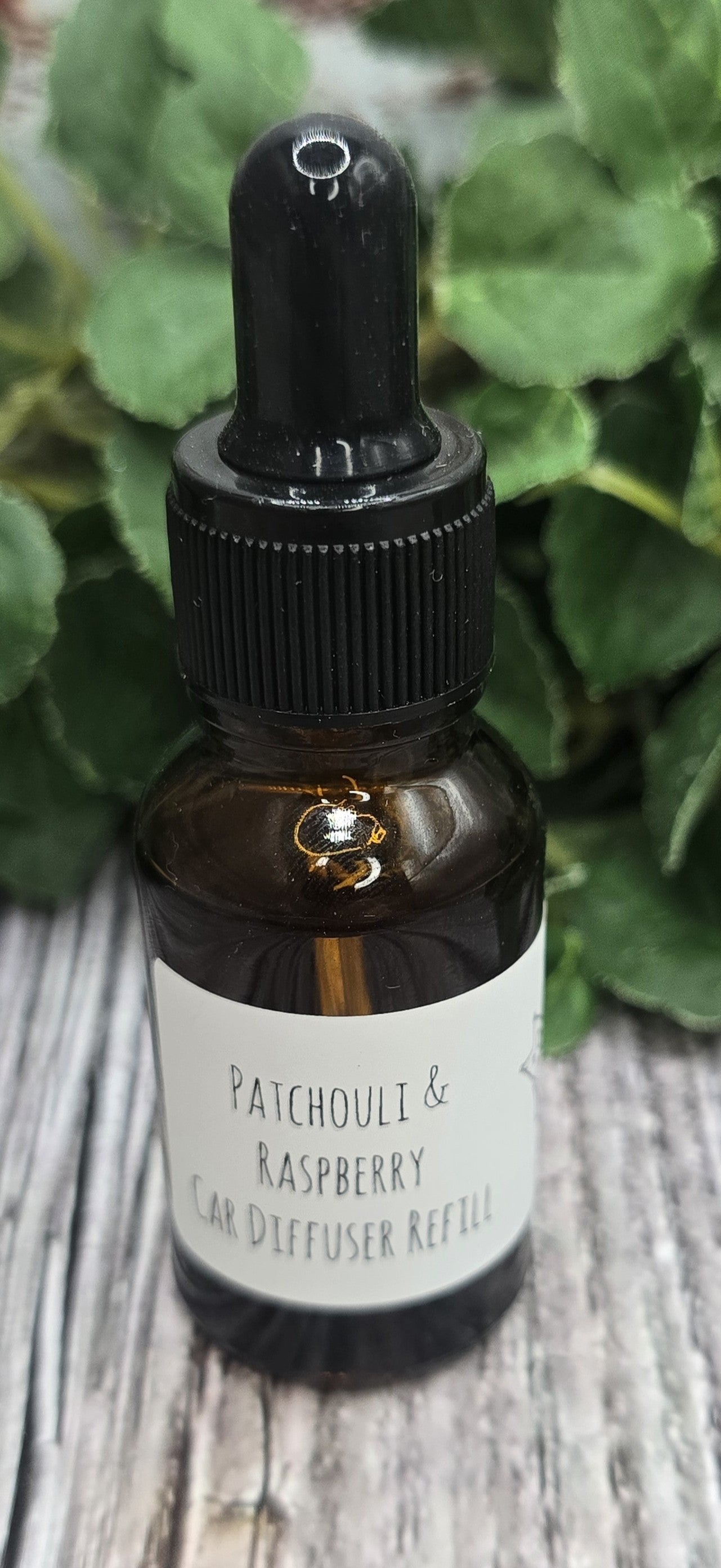 Bottle of Patchouli & Raspberry diffuser refill with dropper on a wooden surface with green leaves in the background
