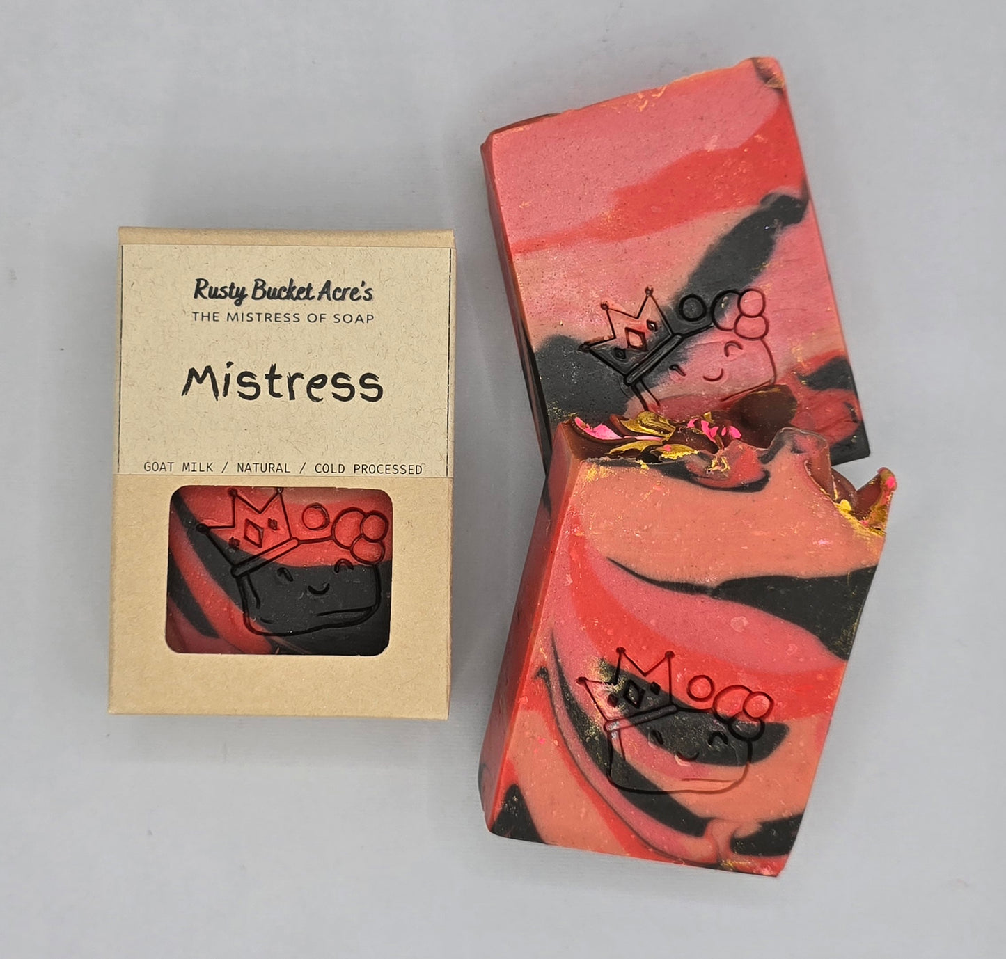 A handmade soap bar with a pink, red, and black swirled pattern and gold foil accents, shown next to its kraft paper packaging box labeled 'Rusty Bucket Acre's The Mistress of Soap Mistress'.
