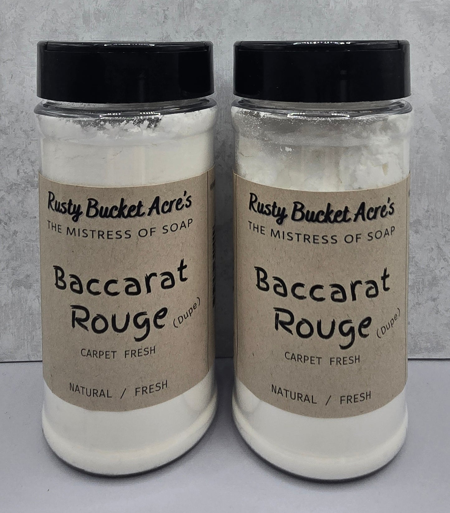 Two clear plastic jars of Rusty Bucket Acre's Baccarat Rouge (Dupe) Carpet Fresh natural fresh soap, filled with white soap powder and capped with black lids, set against a gray textured background.