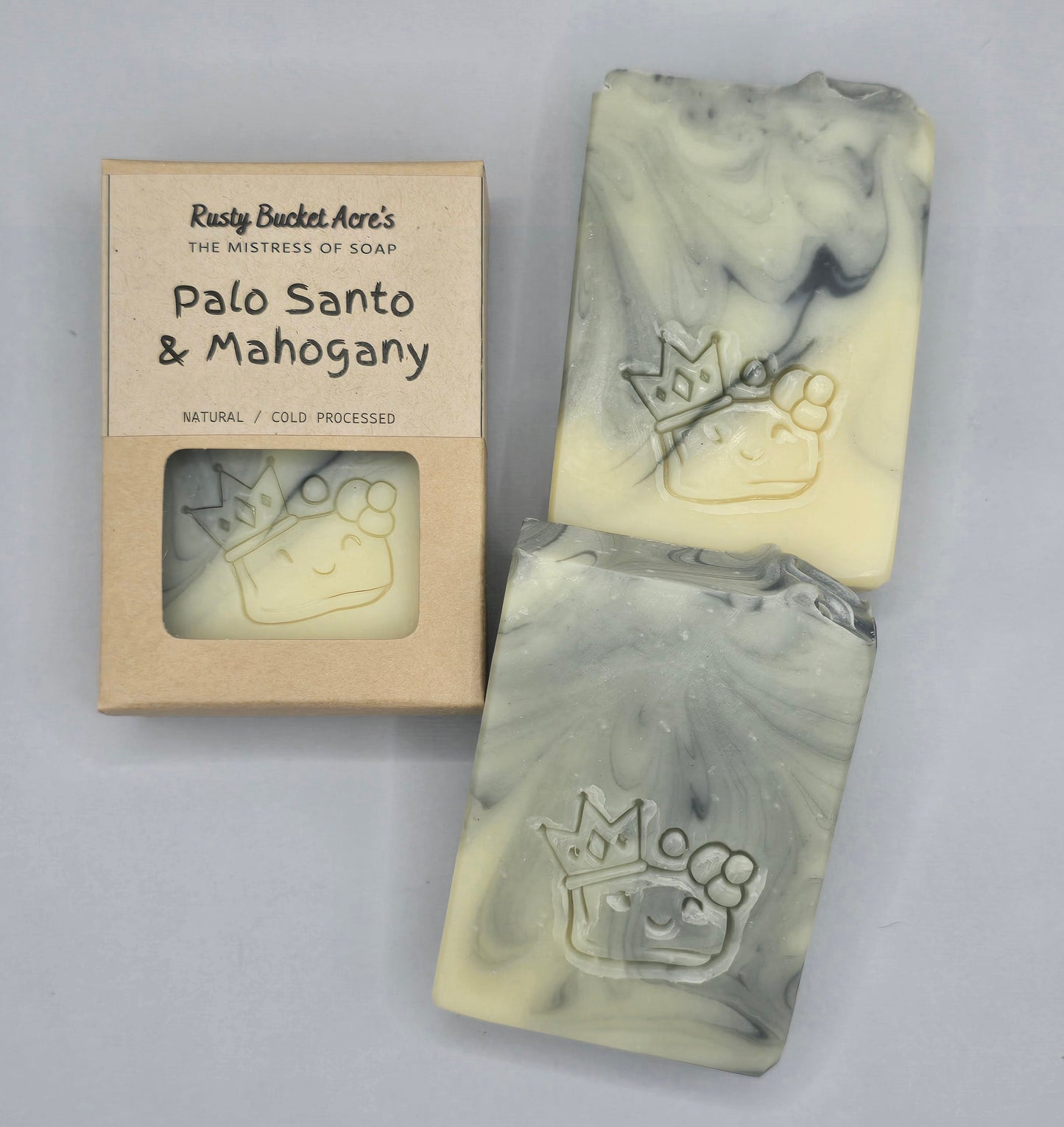 Two handmade cold-pressed soap bars with a cream and gray marbled swirl pattern, embossed with a crown and paw print, displayed next to their kraft paper packaging box labeled 'Rusty Bucket Acre's Palo Santo & Mahogany'.