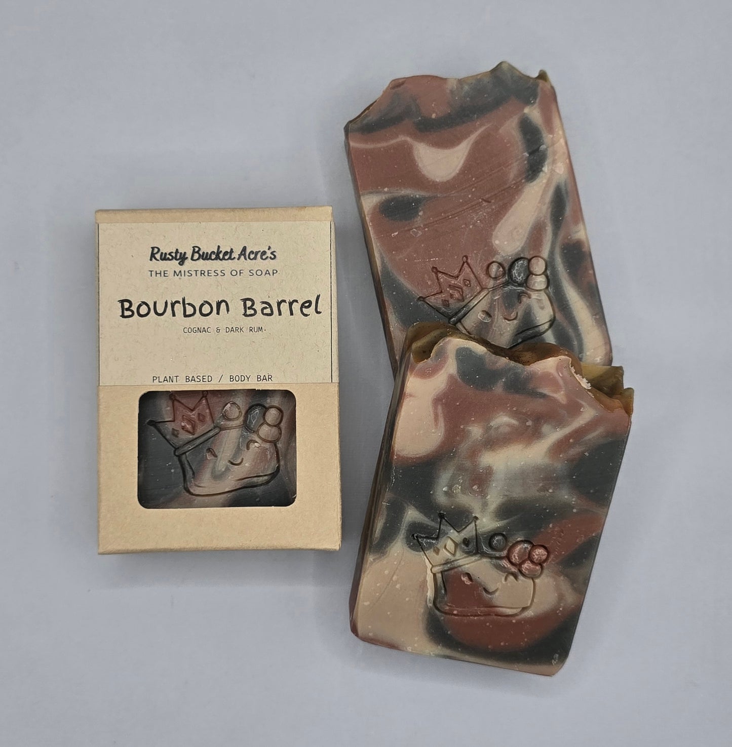 A handmade soap bar with a brown, beige, and black marbled camouflage pattern, shown next to its kraft paper packaging box labeled 'Rusty Bucket Acre's Bourbon Barrel'.