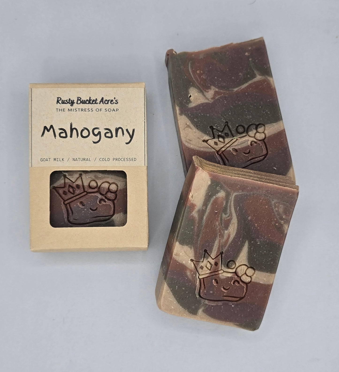 Two bars of handmade soap with a brown and cream marbled pattern, each embossed with a crown and flower design, displayed next to their kraft paper packaging box labeled 'Mahogany' by Rawly Bucket Acre's.