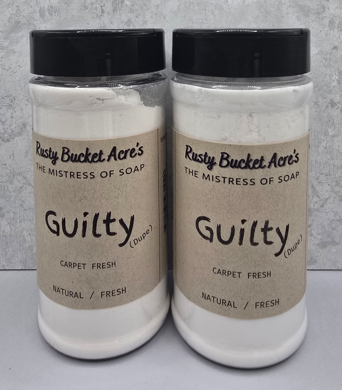 Two clear plastic jars of Rusty Bucket Acre's 'Guilty (Duple)' Carpet Fresh natural soap powder, side-by-side against a gray textured background.