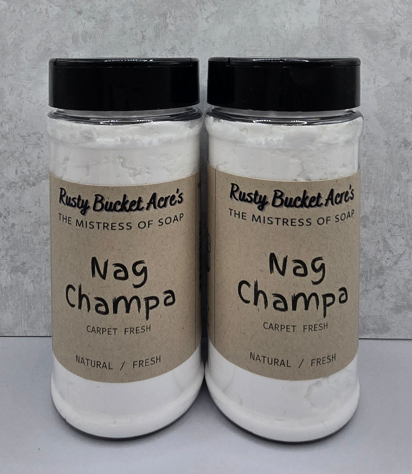 Two clear plastic jars of Rusty Bucket Acre's Nag Champa carpet fresh soap, filled with white soap and topped with black lids, set against a textured gray background.