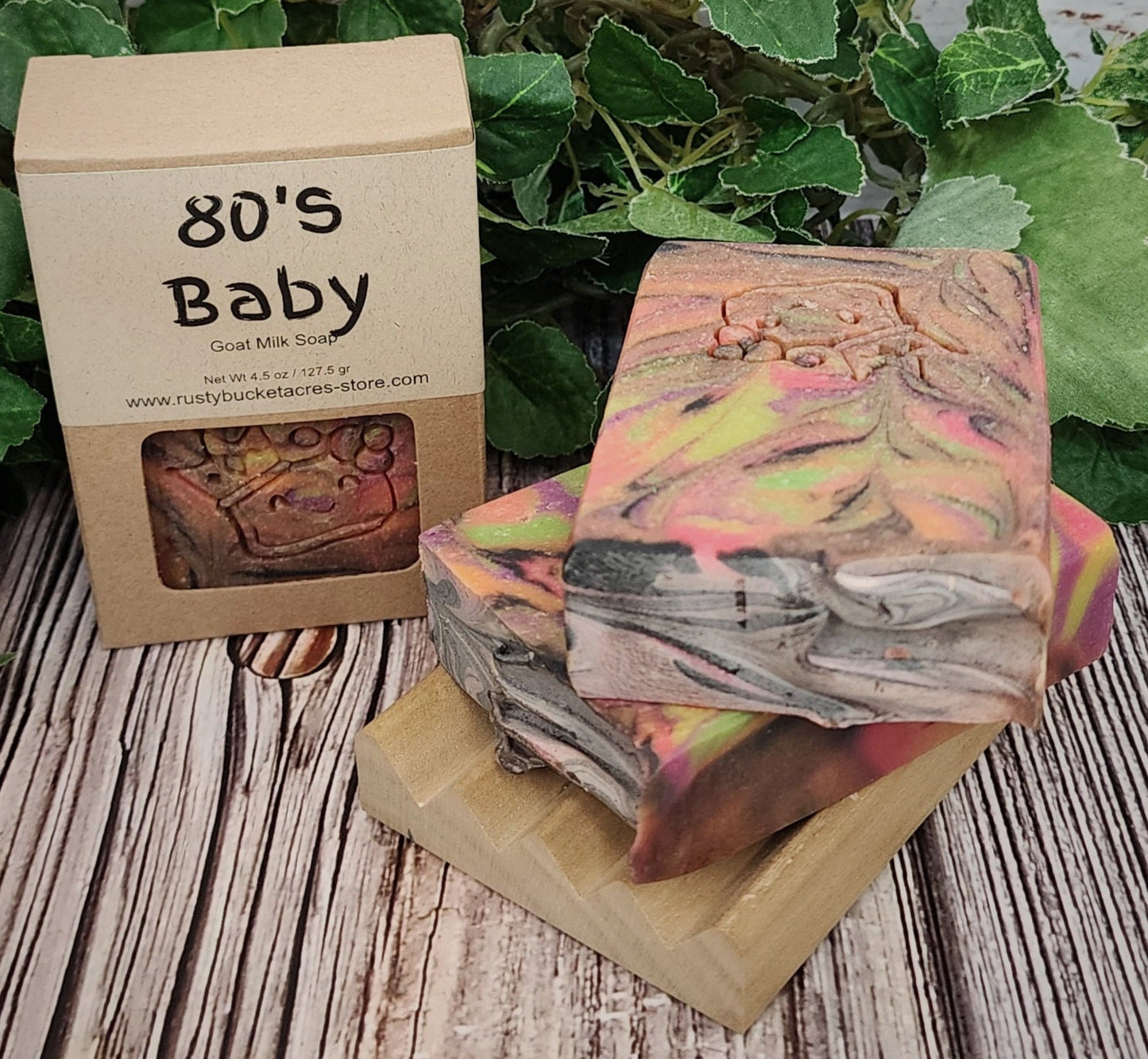Stack of colorful soap bars with a '80's Baby' branded box on a wooden surface with greenery.