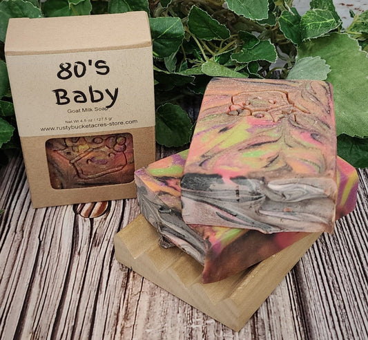 Stack of colorful soap bars with a '80's Baby' branded box on a wooden surface with greenery.