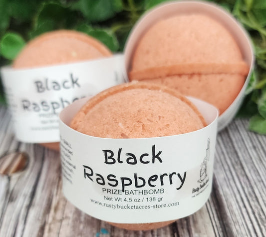 Bath bombs labeled 'Black Raspberry' in white containers on a wooden surface with greenery in the background.