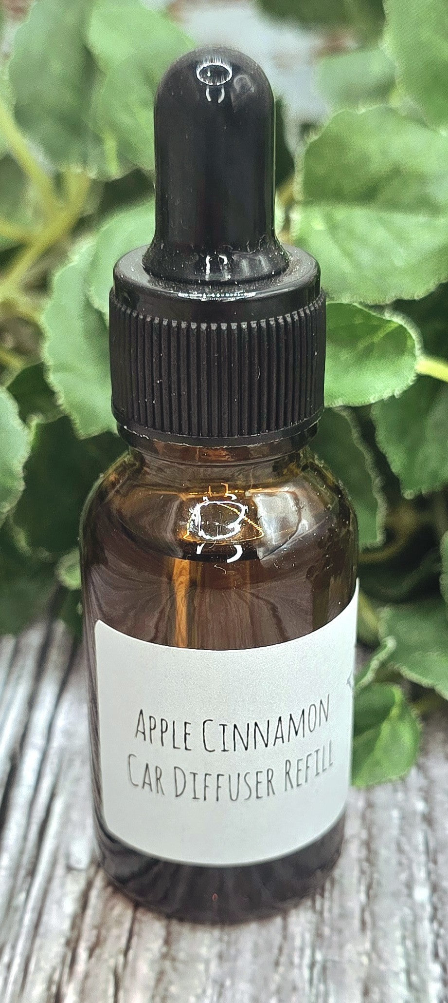 Bottle of Apple Cinnamon Car Diffuser Refill with a dropper on a wooden surface with green leaves in the background