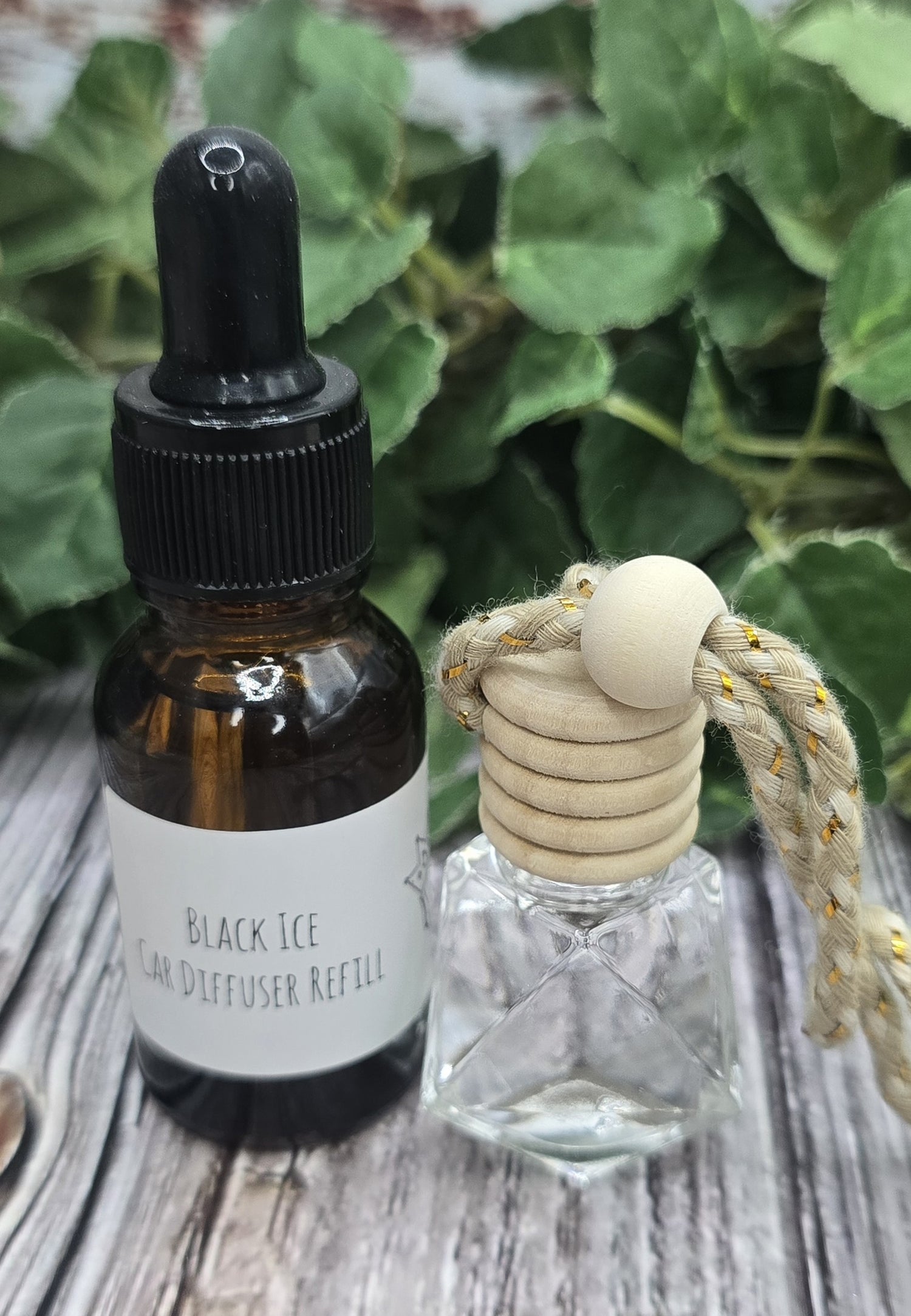 A bottle of 'Black Ice' diffuser oil and a clear glass diffuser with a wooden top on a wooden surface with green leaves in the background.