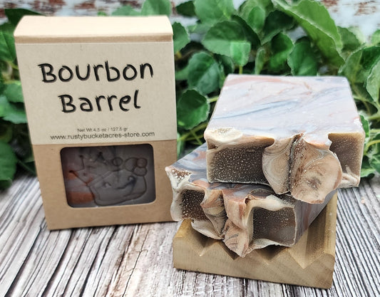 Bar of soap with a decorative top and packaging labeled 'Bourbon Barrel' on a wooden surface with greenery in the background.