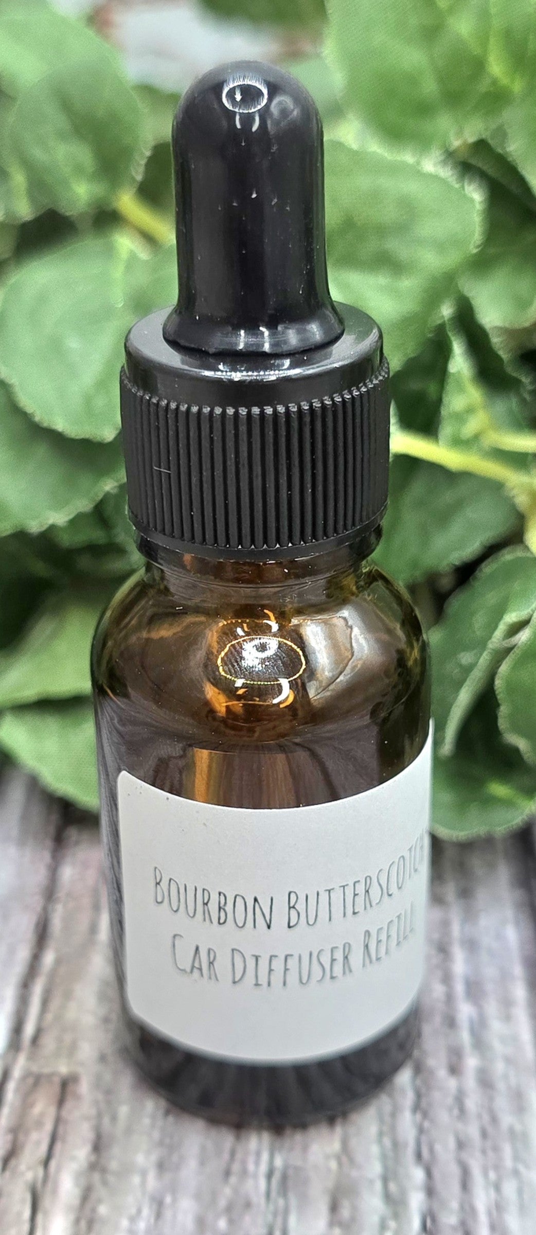 Bottle of Bourbon Butterscotch Car Diffuser Refill with a dropper on a wooden surface with green leaves in the background