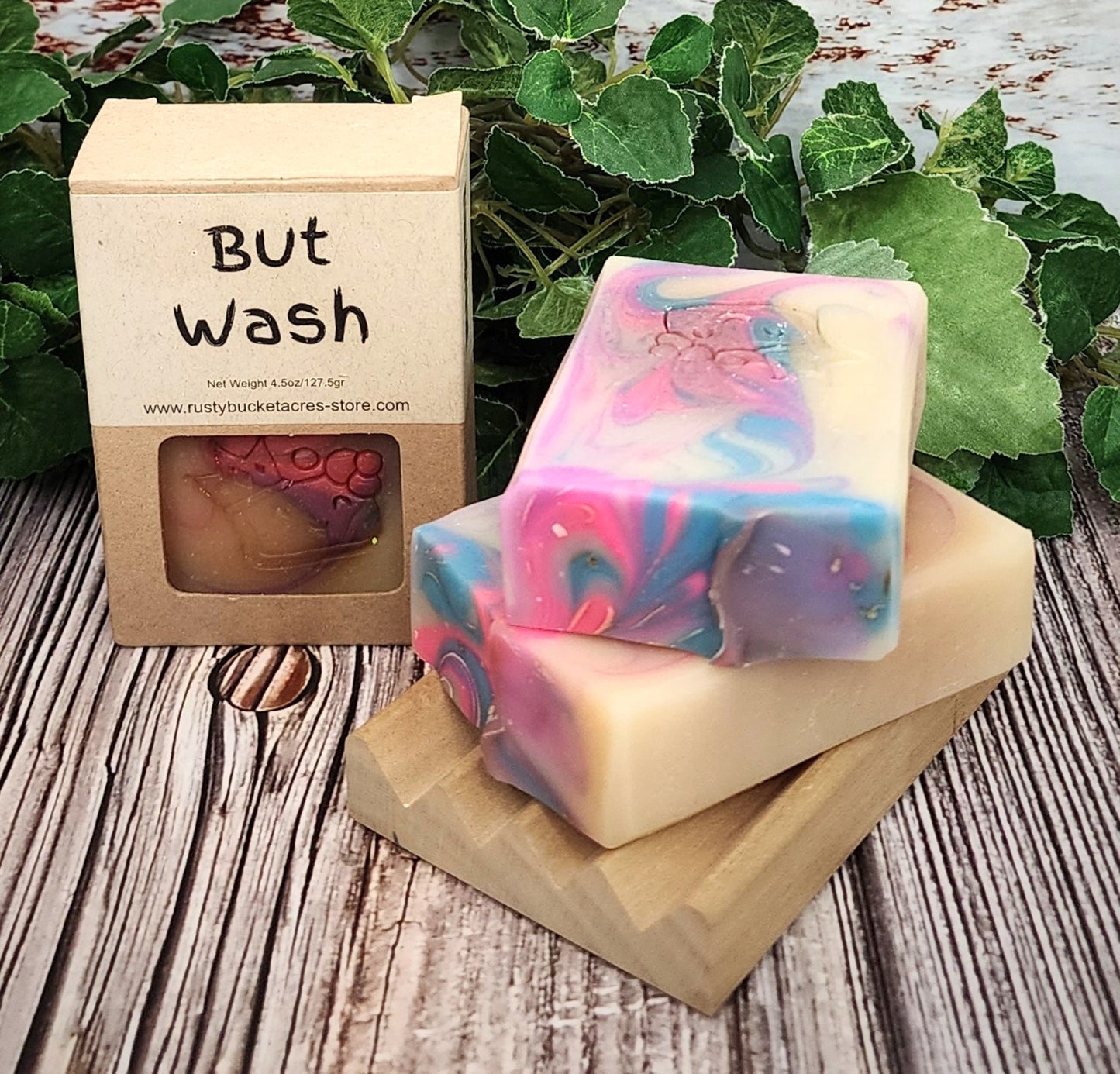Colorful soap bar with a wooden box labeled 'But Wash' on a wooden surface with green leaves in the background.