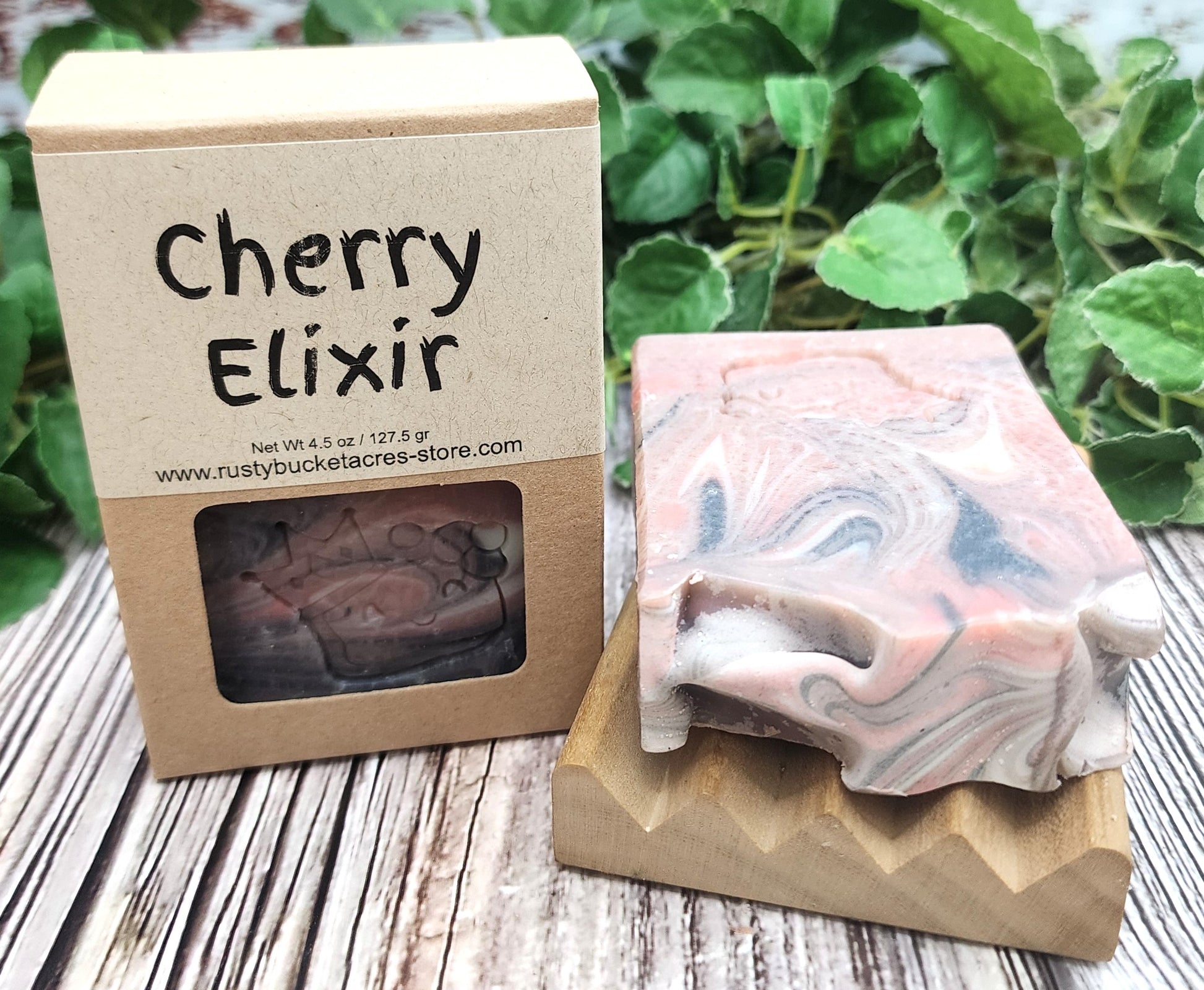 Bar of soap with 'Cherry Elixir' packaging on a wooden surface with greenery in the background