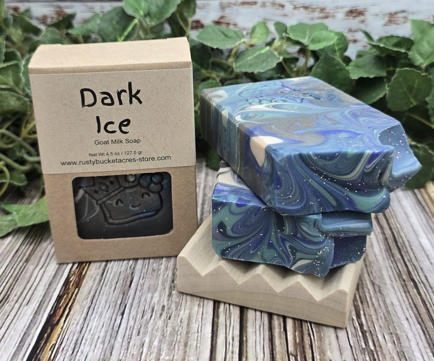 Dark Ice goat milk soap with packaging on a wooden surface with greenery in the background