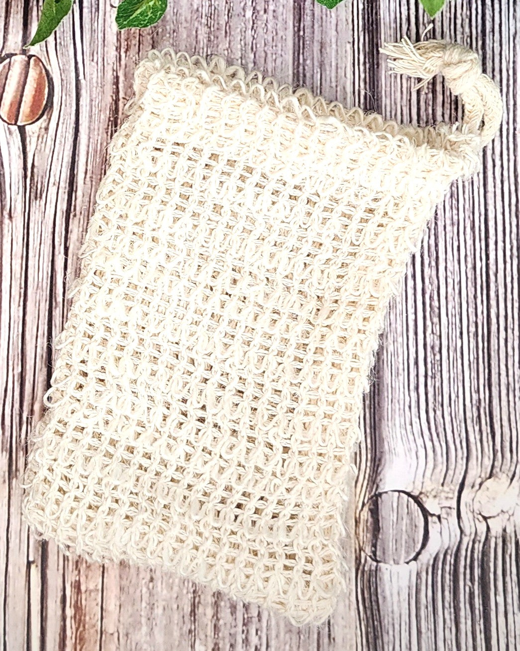 Beige woven soap bag on a wooden surface with green leaves and a spoon.