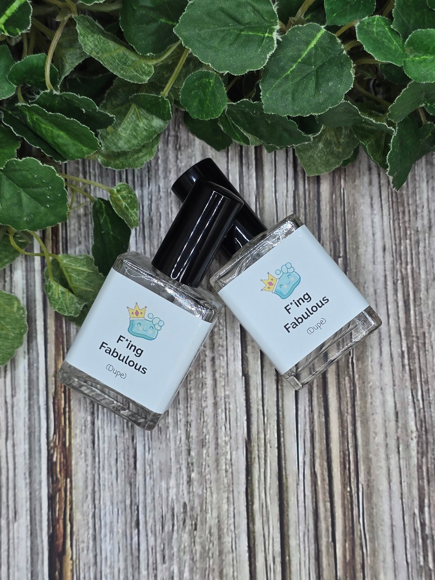 Two small bottles of perfume labeled 'Fling Fabulous' on a wooden surface with green leaves.