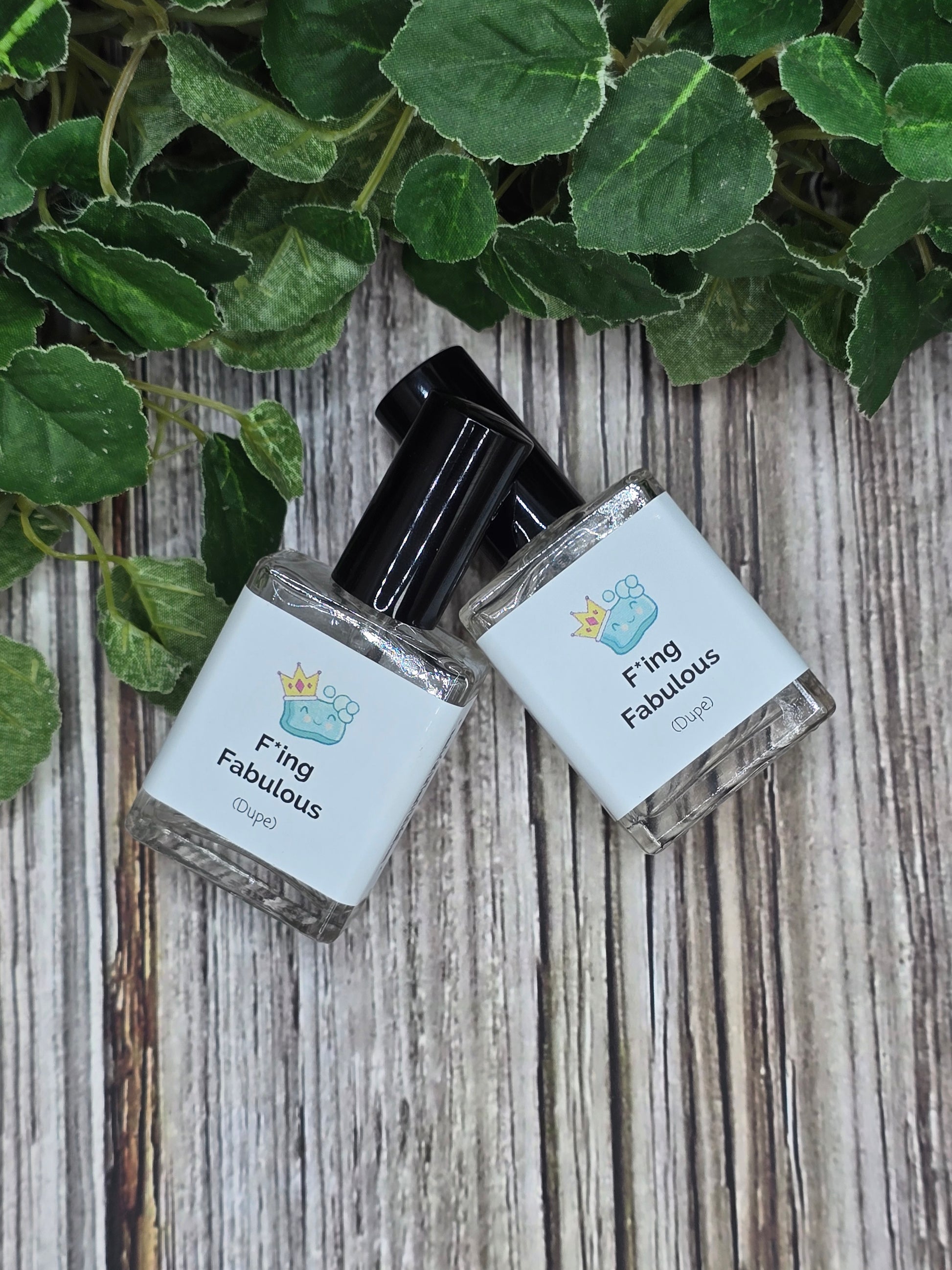 Two small bottles of perfume labeled 'Fling Fabulous' on a wooden surface with green leaves.