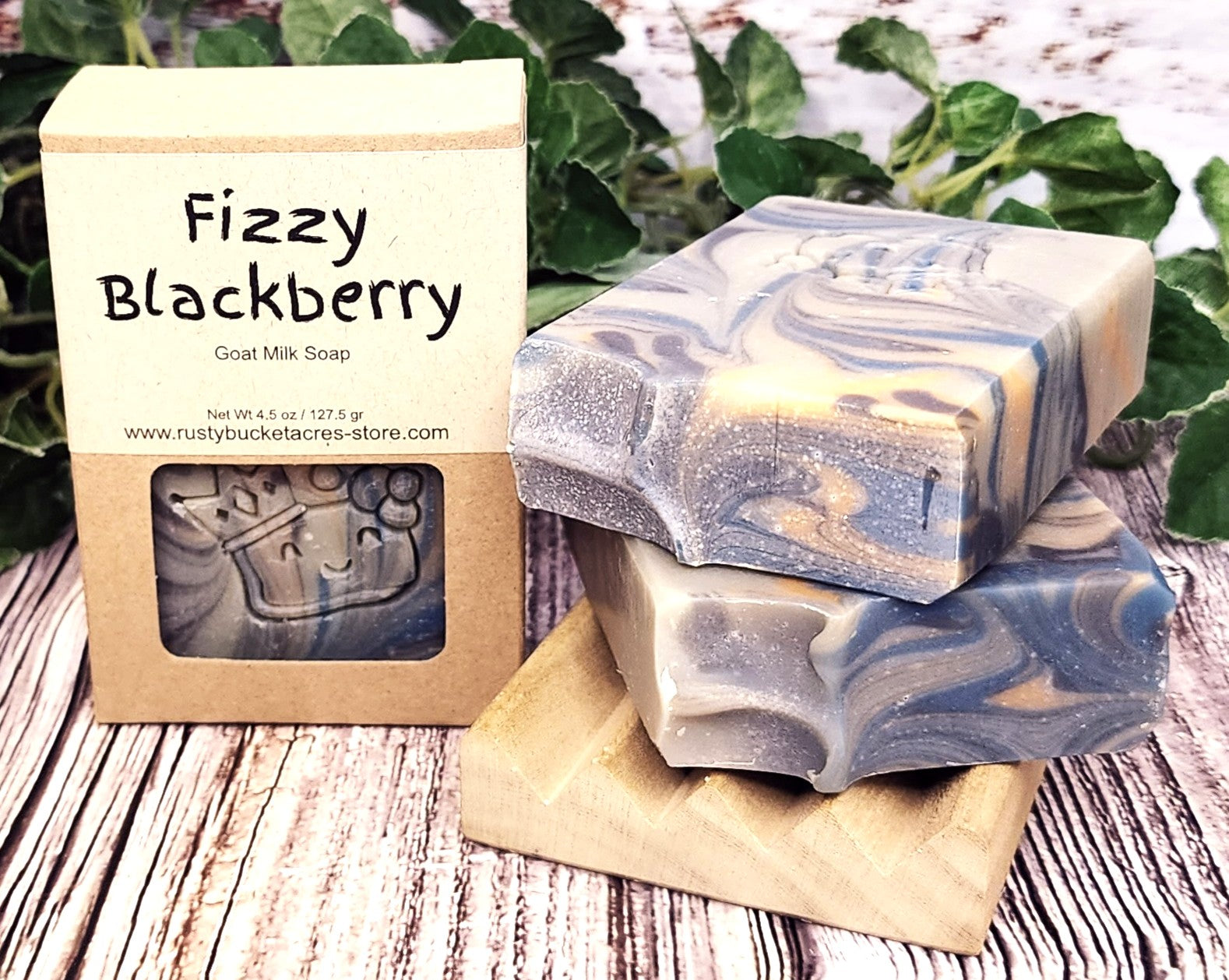 Fizzy Blackberry goat milk soap with packaging on a wooden surface with greenery in the background