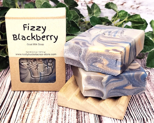 Fizzy Blackberry goat milk soap with packaging on a wooden surface with greenery in the background