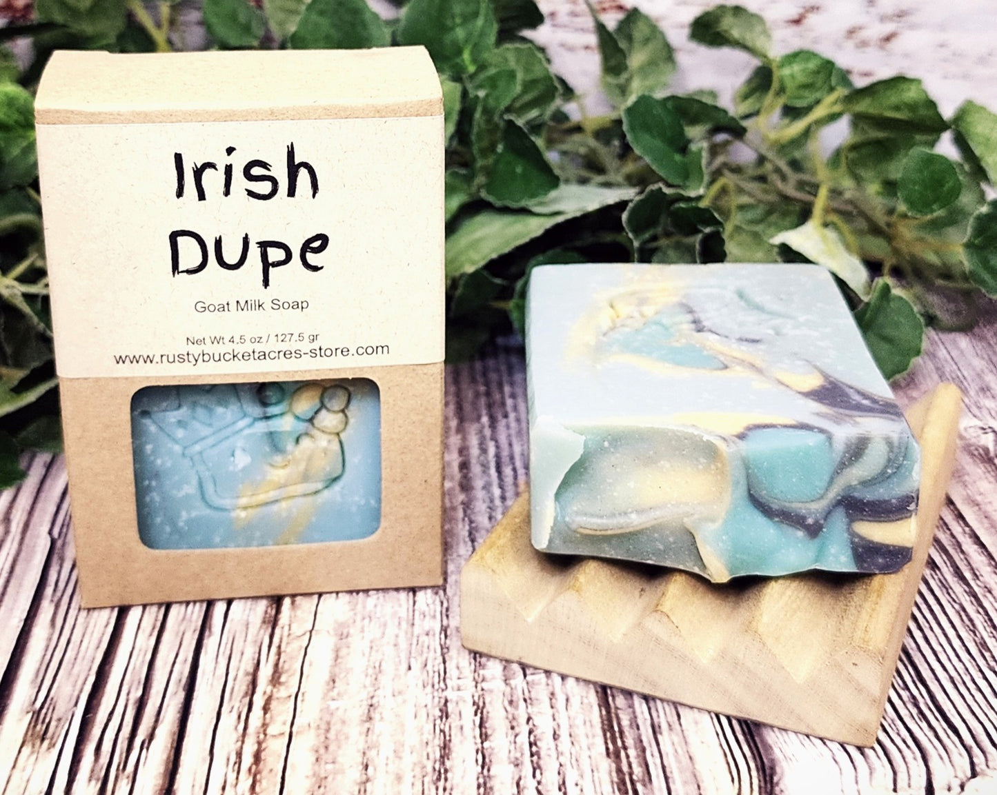 Irish Dupe goat milk soap with packaging on a wooden surface with greenery in the background
