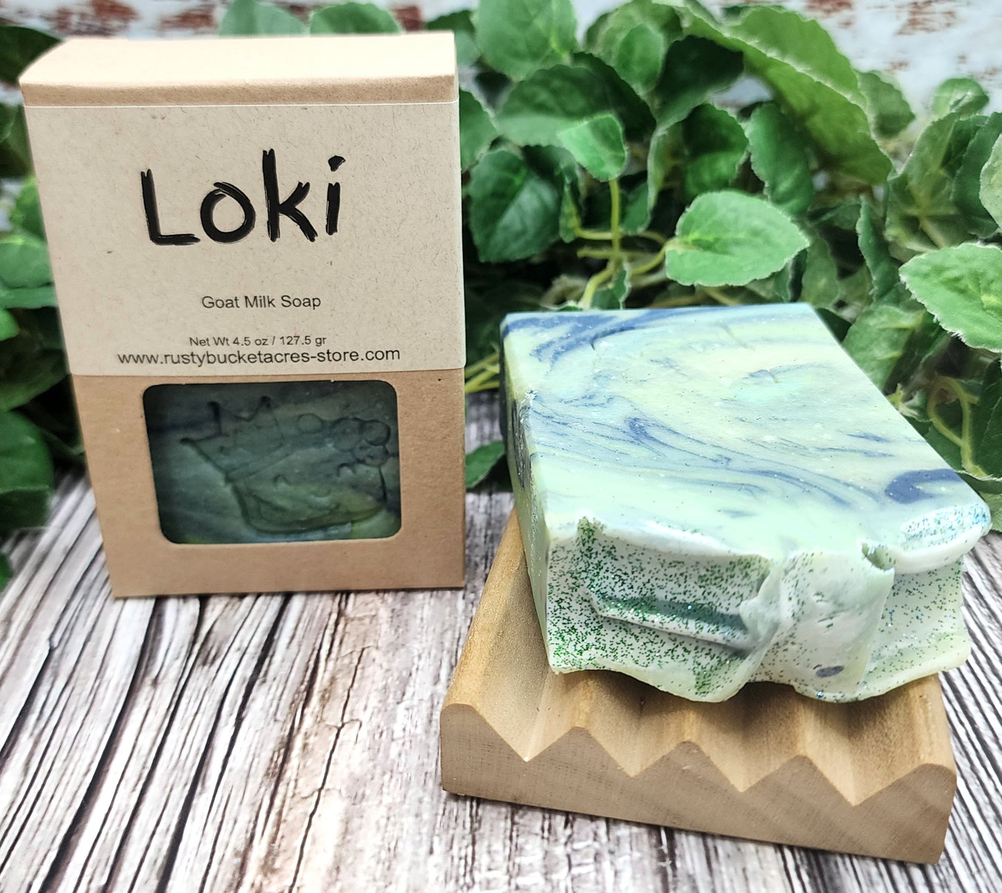Goat milk soap bar with 'Loki' branding on a wooden stand and packaging, surrounded by greenery.