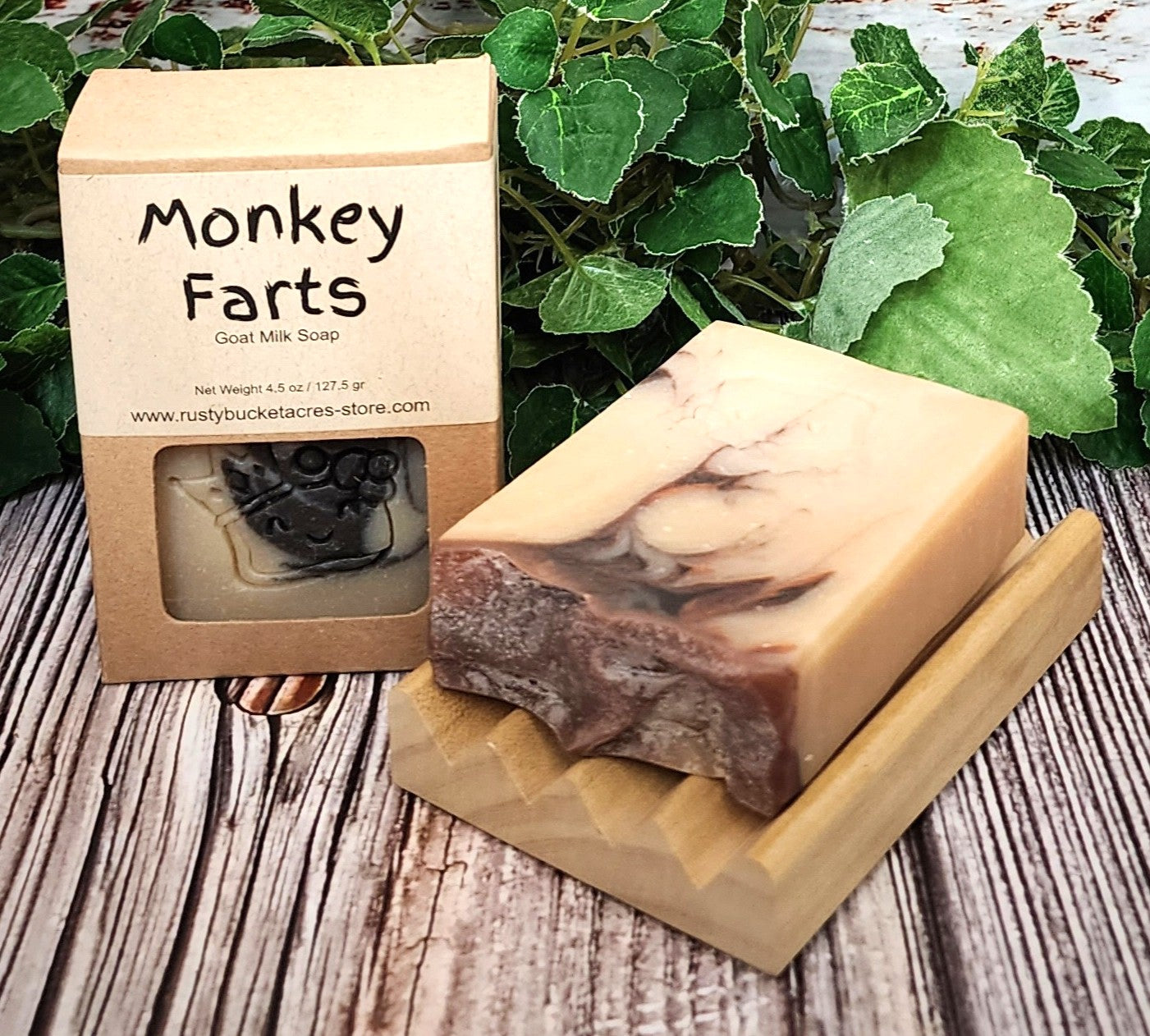 Goat milk soap with a marbled design on a wooden block, next to its packaging labeled 'Monkey Farts' on a wooden surface with green leaves in the background.