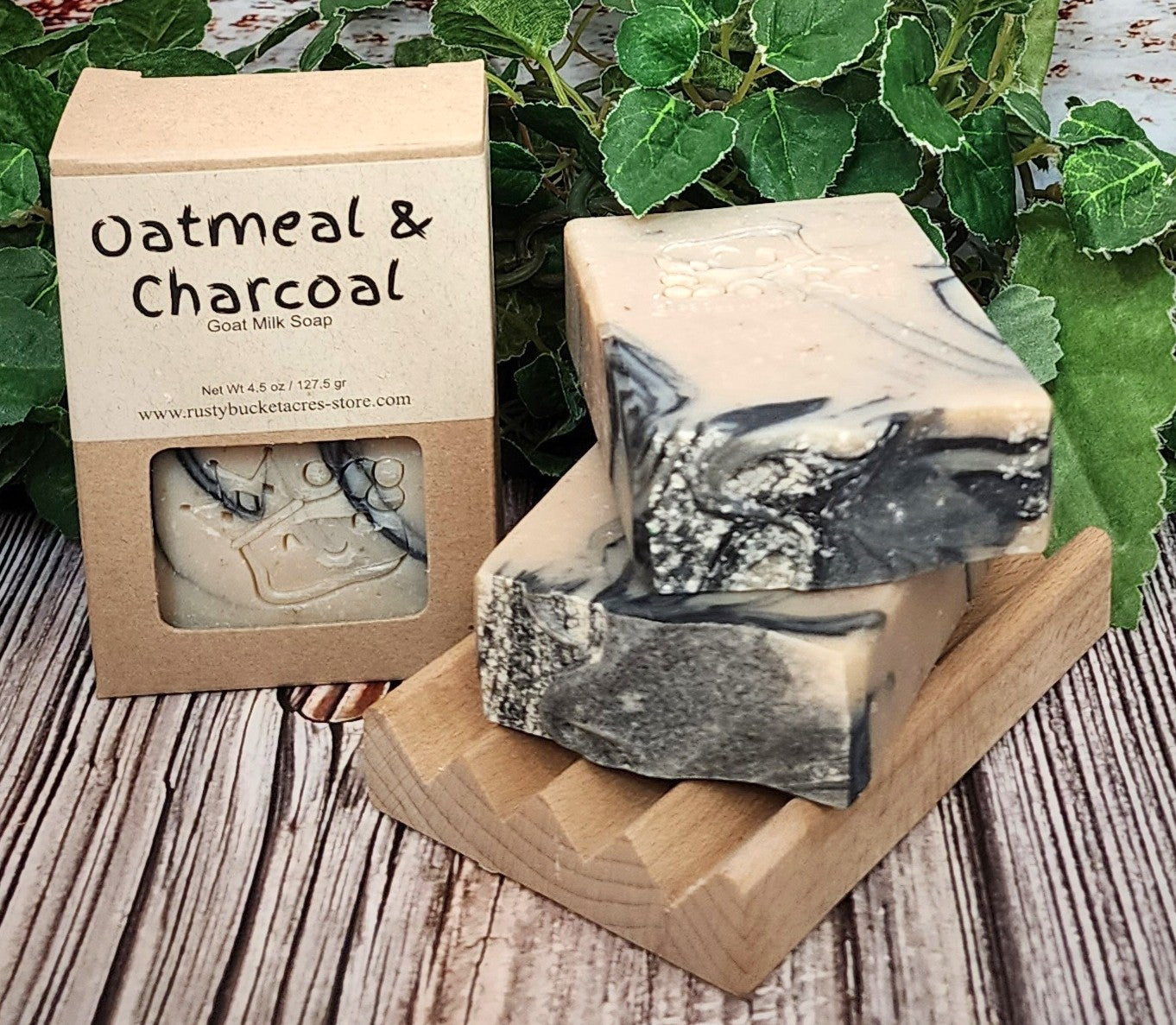 Oatmeal & Charcoal soap bars with packaging on a wooden surface with greenery in the background