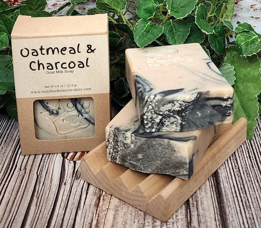 Oatmeal & Charcoal soap bars with packaging on a wooden surface with greenery in the background