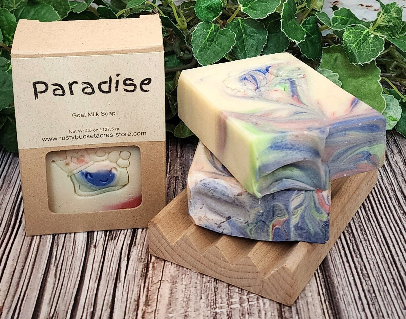 Colorful soap bars with a 'Paradise' branded box on a wooden surface with greenery in the background.