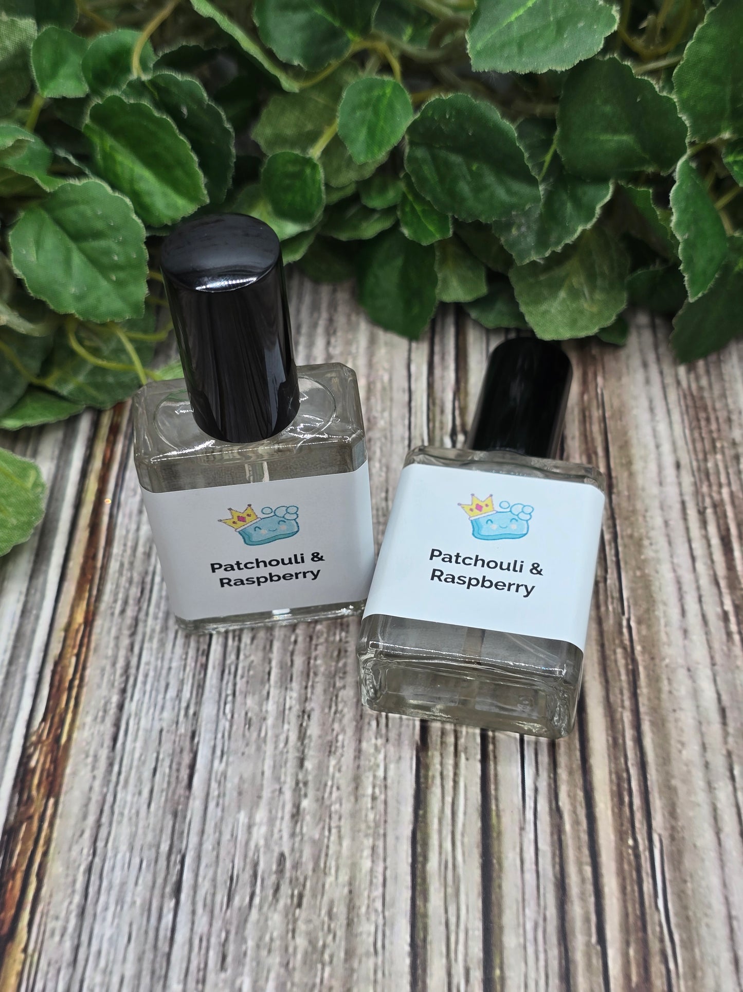 Two small bottles of Patchouli Raspberry perfume on a wooden surface with green leaves.