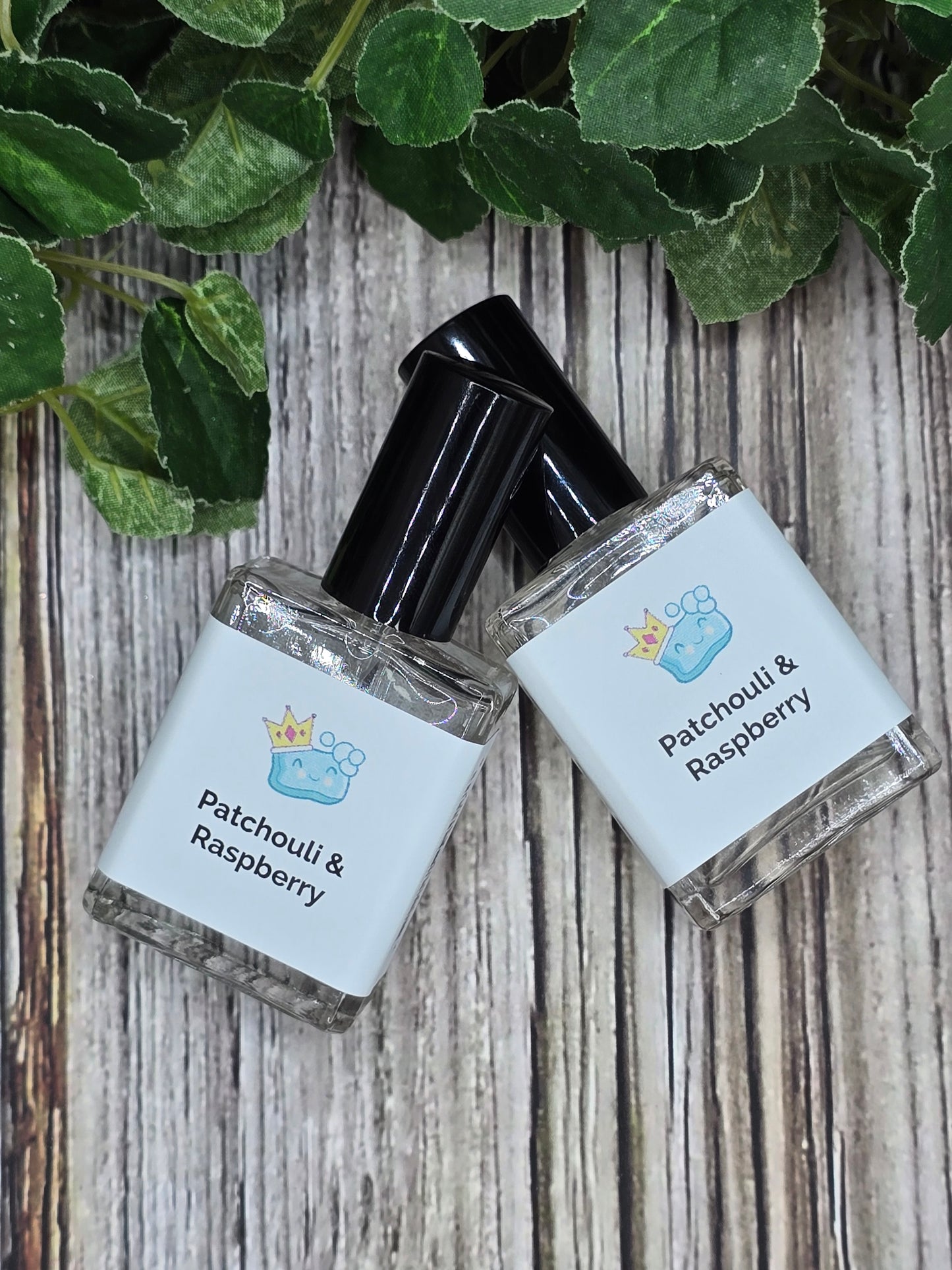 Two small bottles of perfume labeled 'Patchouli & Raspberry' on a wooden surface with green leaves.
