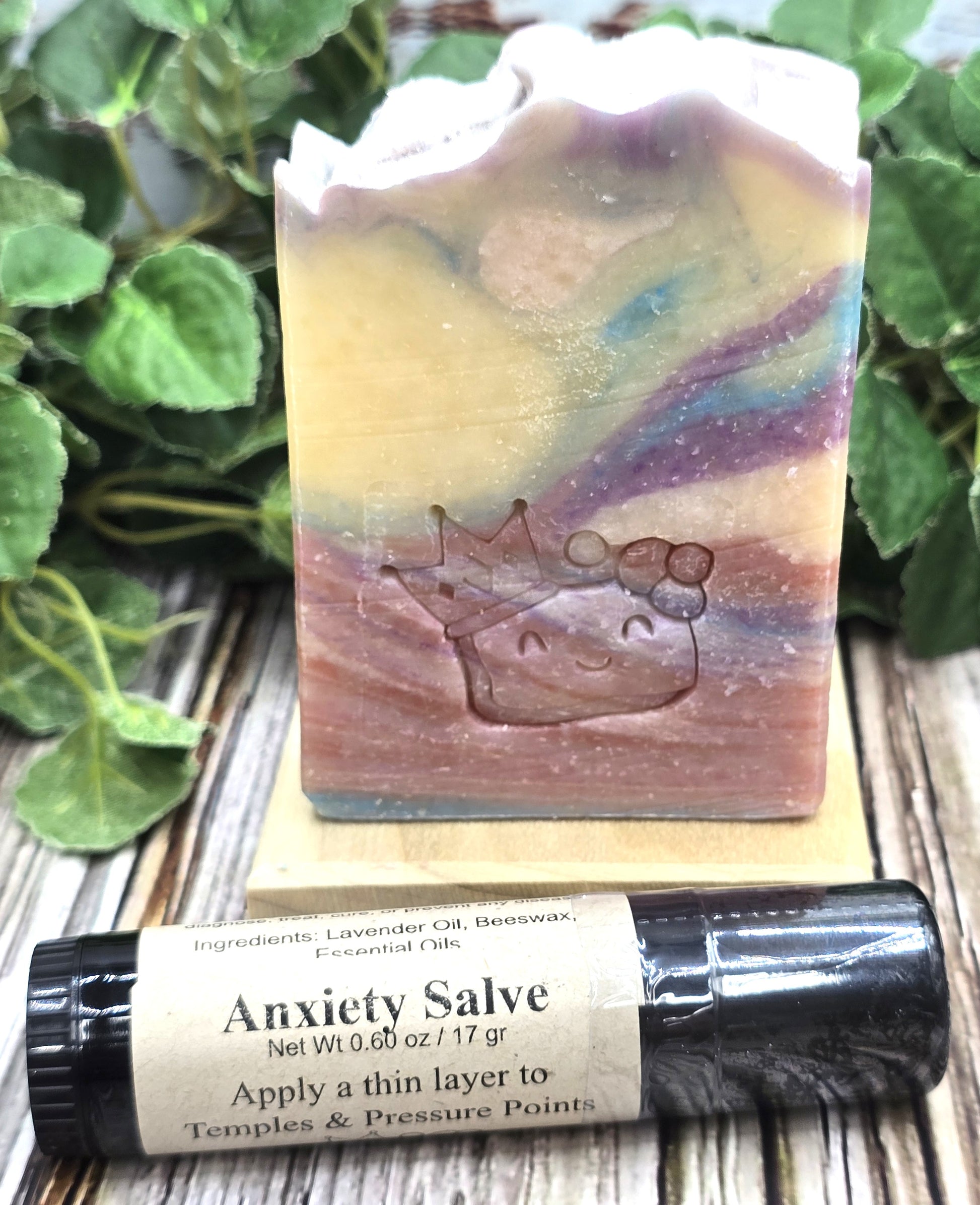 Colorful soap bar with a crown design and Anxiety Salve twist up tube on a wooden surface with greenery.