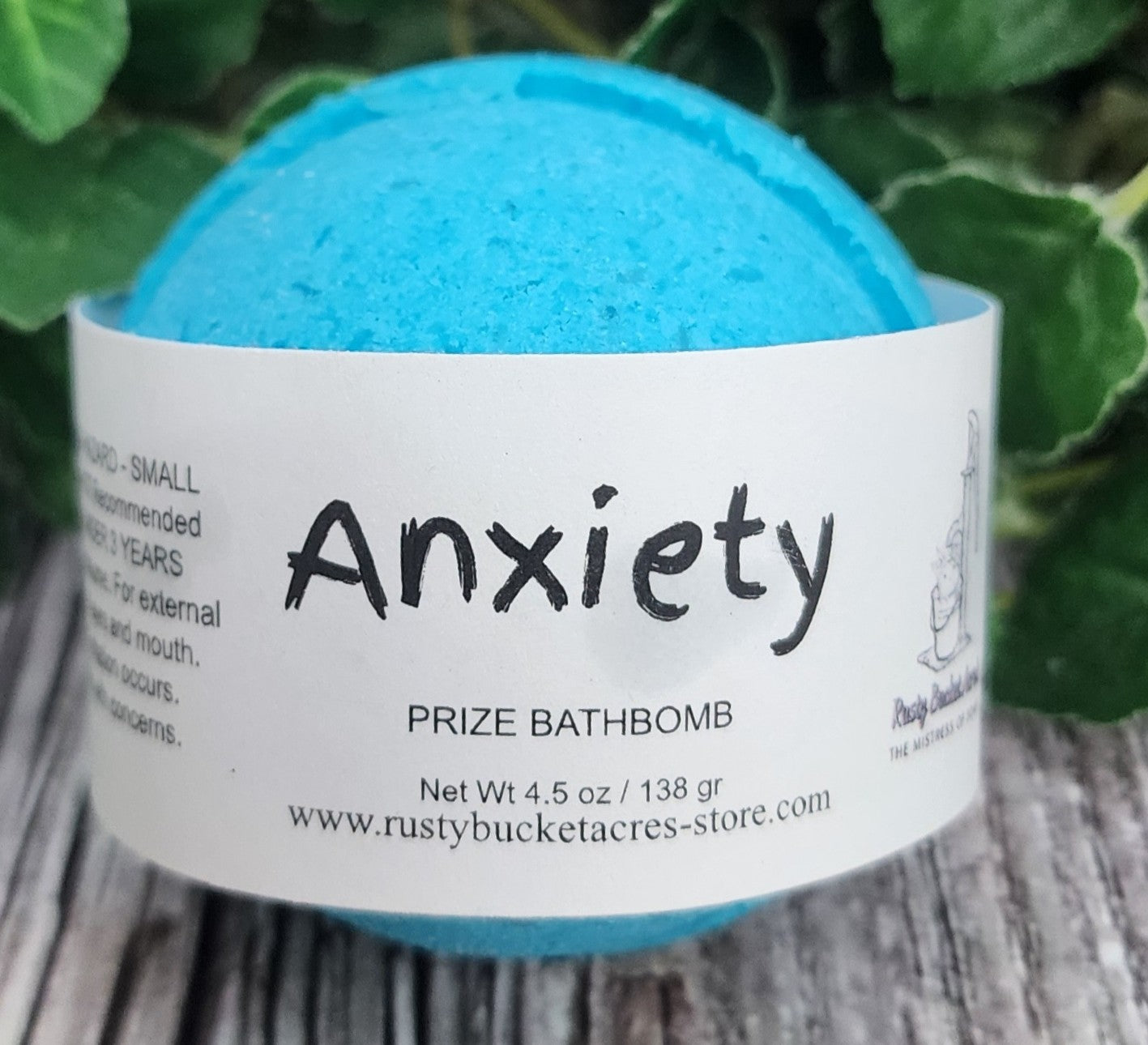 Blue bath bomb labeled 'Anxiety' with a white label on a wooden surface and green leaves in the background.