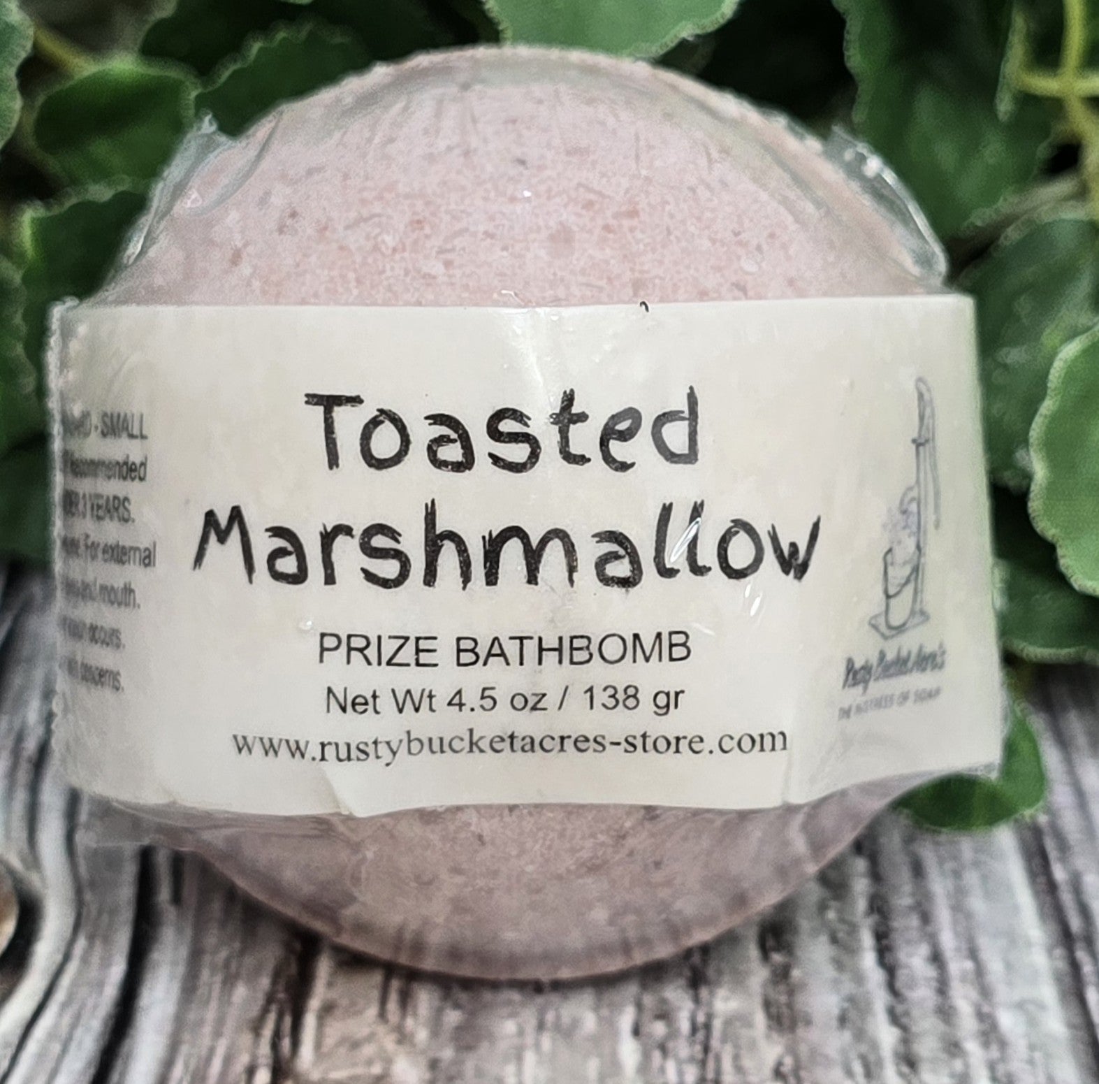 Bath bomb labeled 'Toasted Marshmallow' on a wooden surface with greenery in the background