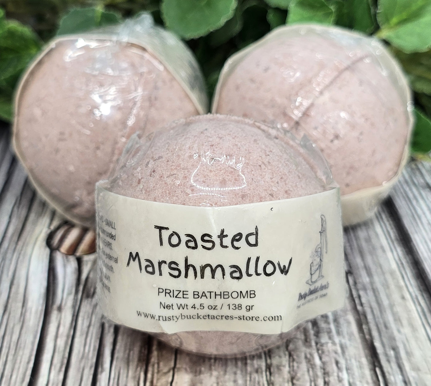 Bath bomb labeled 'Toasted Marshmallow' on a wooden surface with greenery in the background