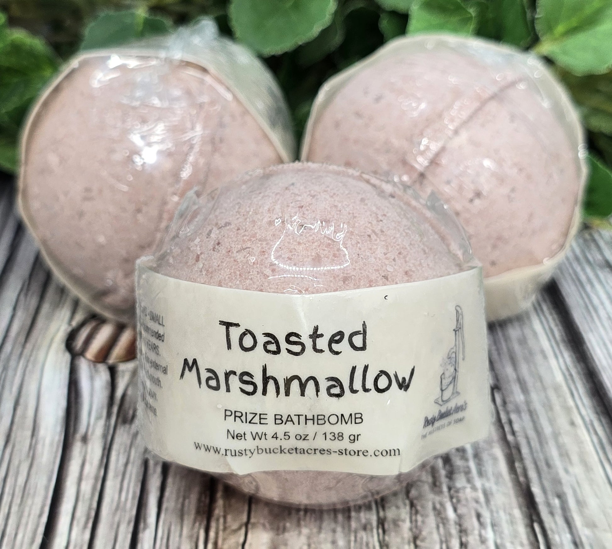 Bath bomb labeled 'Toasted Marshmallow' on a wooden surface with greenery in the background