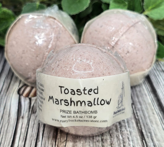 Bath bomb labeled 'Toasted Marshmallow' on a wooden surface with greenery in the background