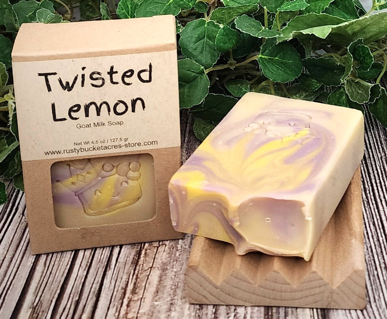Goat milk soap bar with 'Twisted Lemon' packaging on a wooden surface with green leaves in the background.