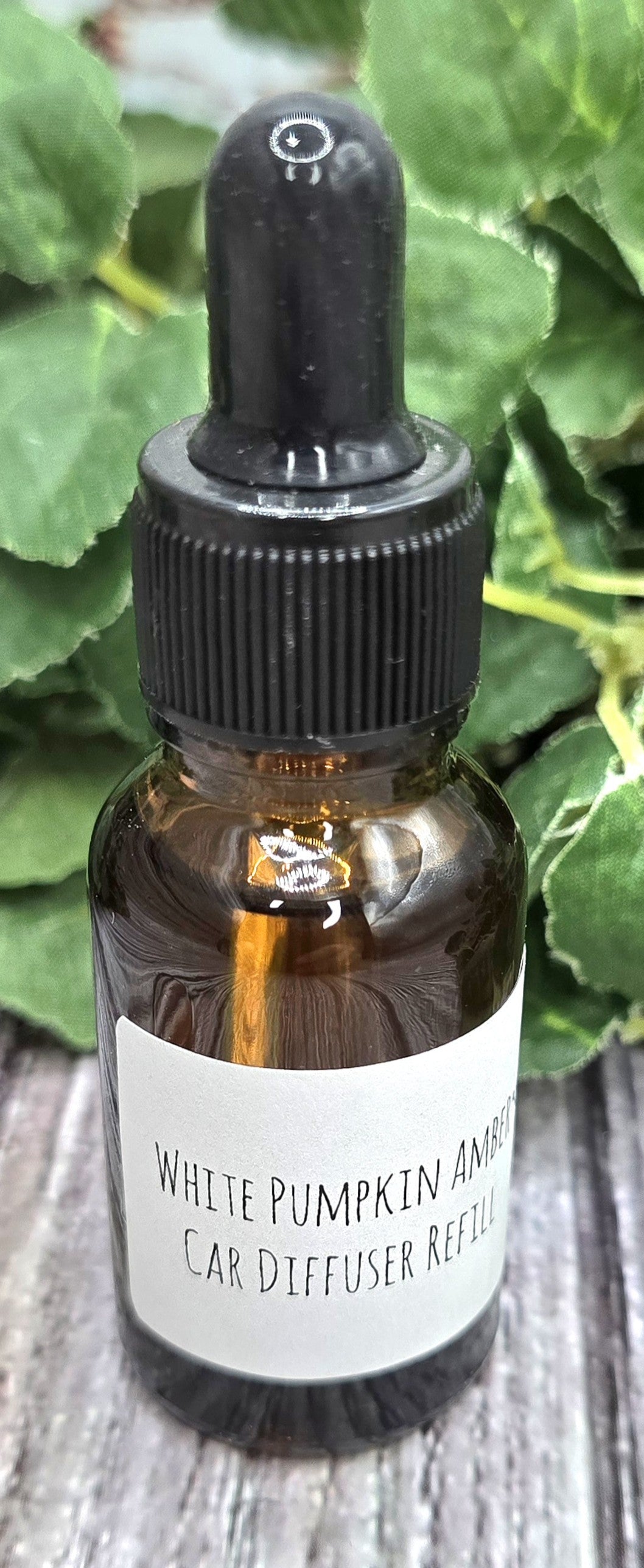 Brown glass bottle with dropper labeled 'White Pumpkin Amber' car diffuser refill on a wooden surface with green leaves in the background.