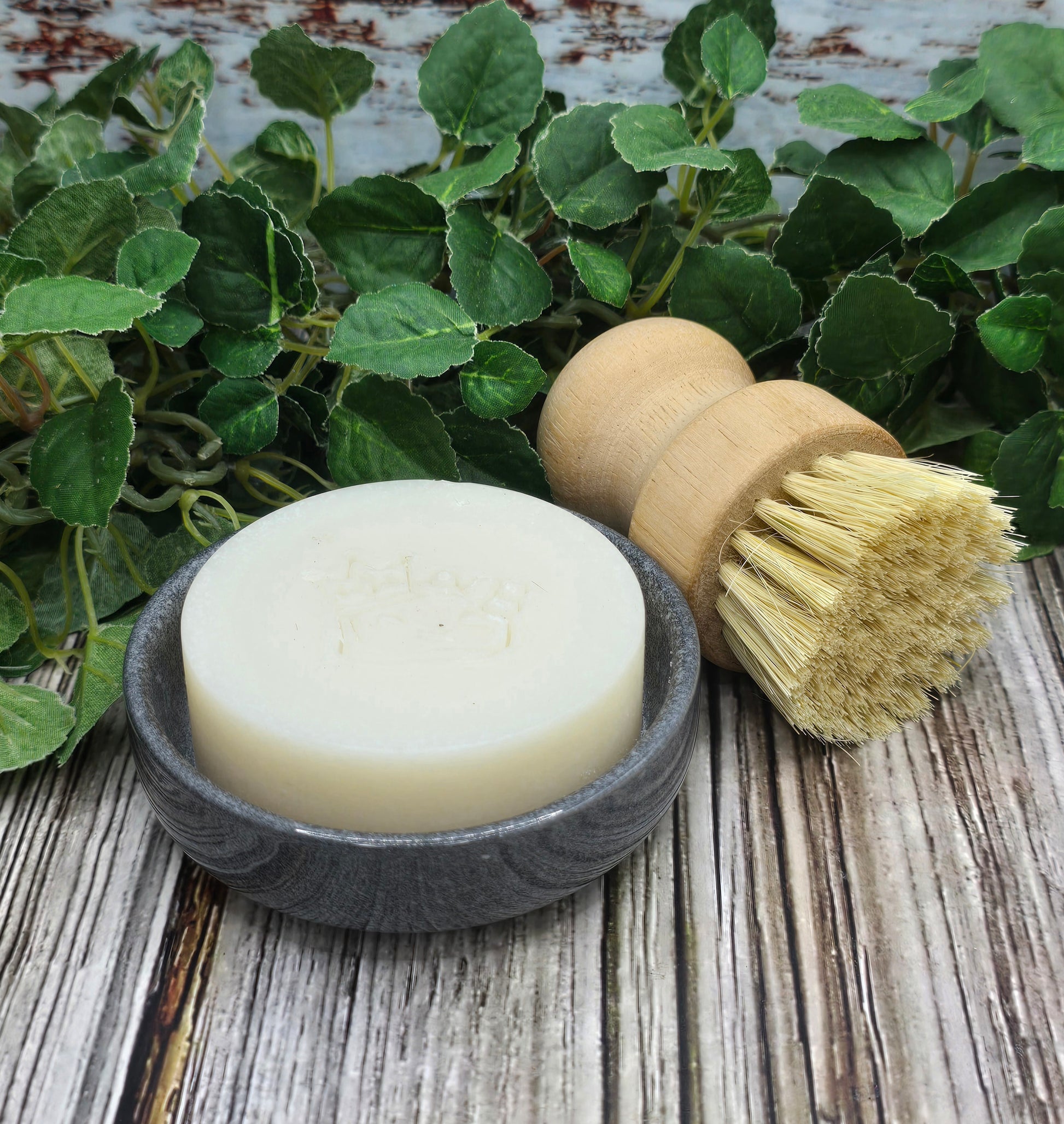Bar of soap in a stone dish with a wooden brush on a wooden surface with green leaves.