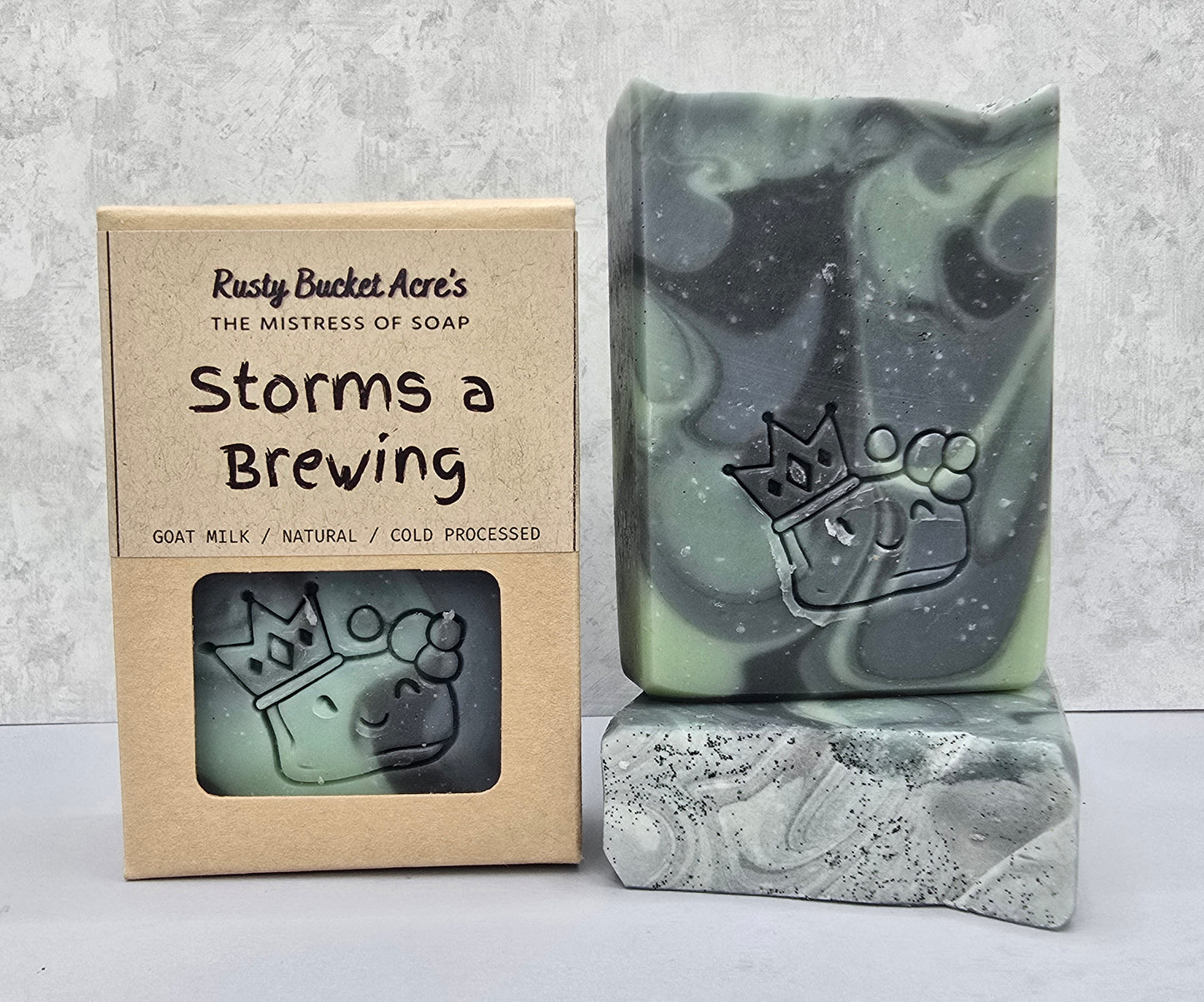 A handmade soap bar with a swirled green, black, and gray marbled pattern and an embossed paw print with a crown, displayed next to its kraft paper packaging box labeled 'Storms a Brewing' by Rusty Bucket Acre's.