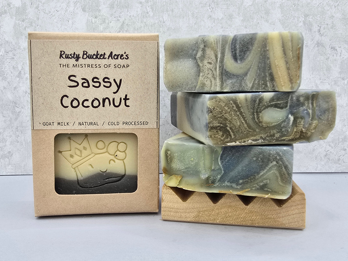 Three marbled handmade soap bars with green, gray, and cream swirls are stacked on a small wooden stand next to their kraft paper packaging box labeled 'Rusty Bucket Acre's Sassy Coconut'.