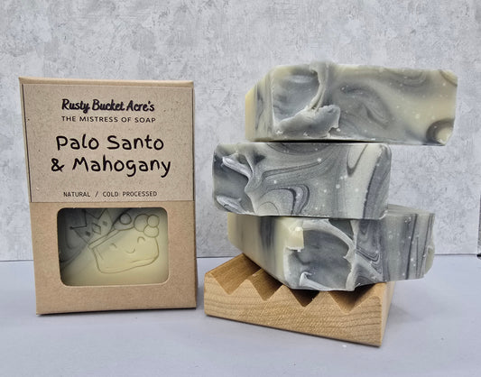Three marbled gray and off-white soap bars displayed on a small wooden stand next to their kraft paper packaging box labeled 'Rusty Bucket Acre's Palo Santo & Mahogany'.