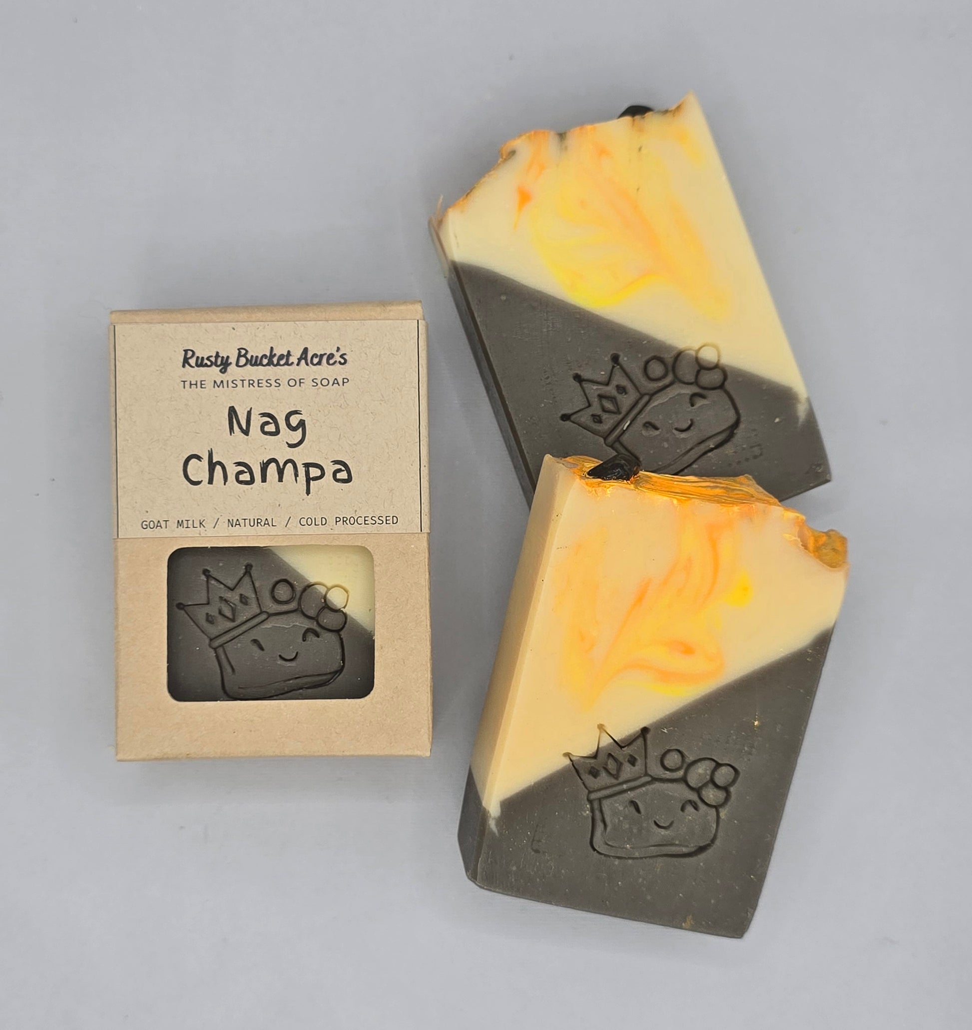 Two bars of Rusty Bucket Acre's Nag Champa goat milk soap with a yellow and orange swirled pattern on a dark gray base, shown next to their kraft paper packaging box on a light gray background.