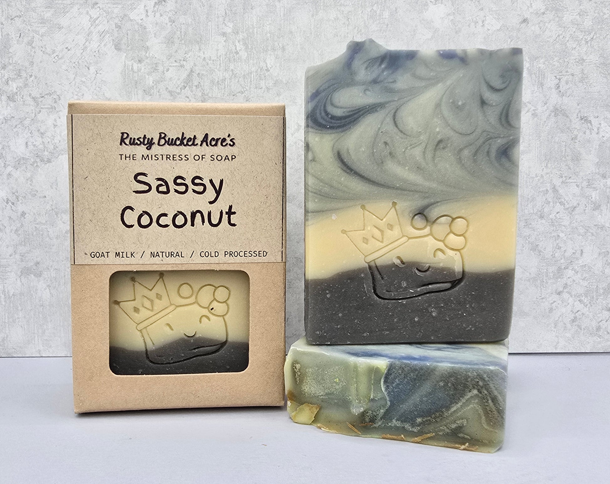 A handmade soap bar with a swirled blue, cream, and dark gray pattern, embossed with a crown and coconut design, shown next to its kraft paper packaging box labeled 'Sassy Coconut' by Rusty Bucket Aloe's.