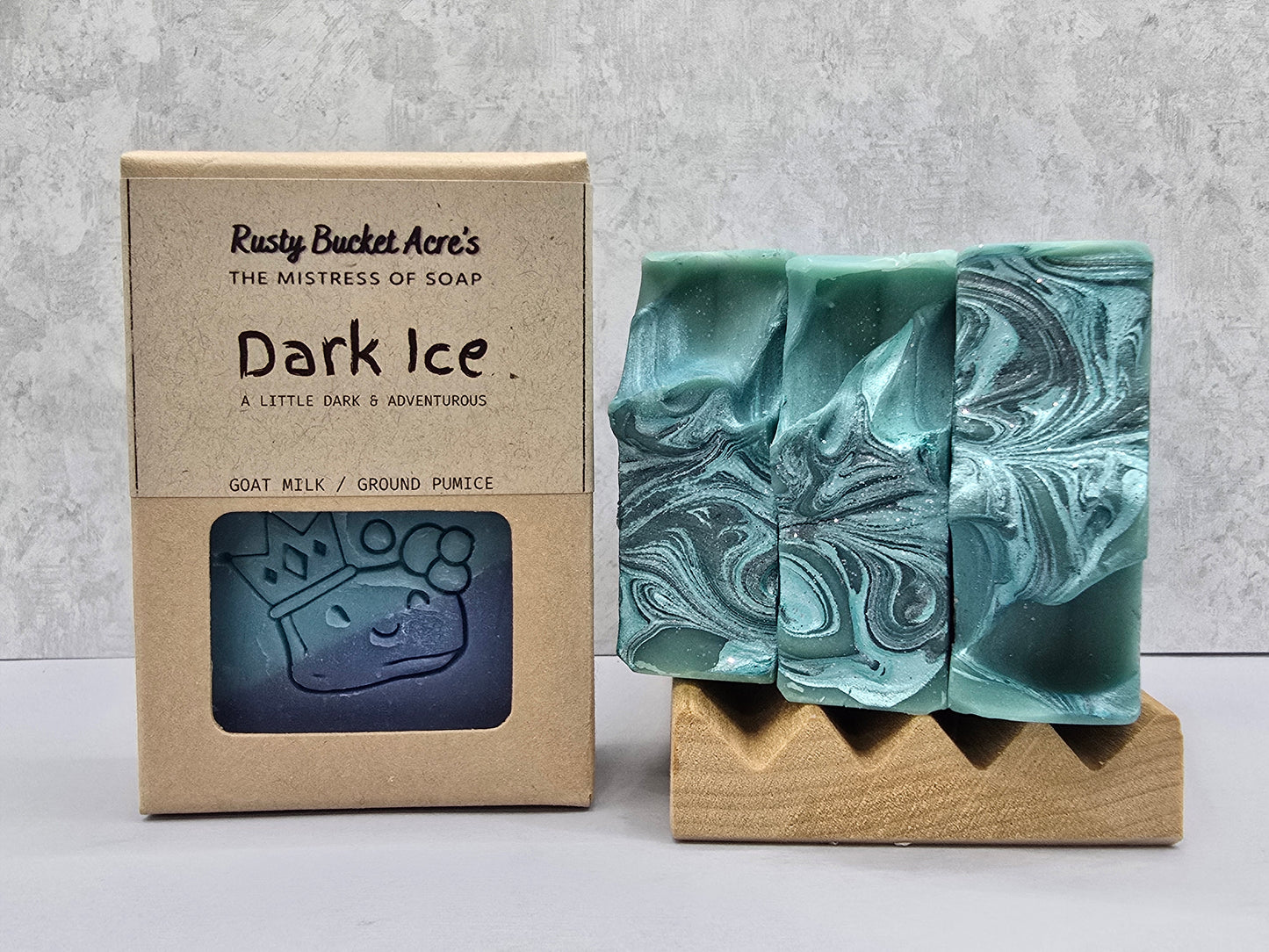A handmade soap bar with a swirled teal and white pattern displayed on a wooden stand next to its kraft paper packaging box labeled 'Rusty Bucket Acre's Dark Ice'.