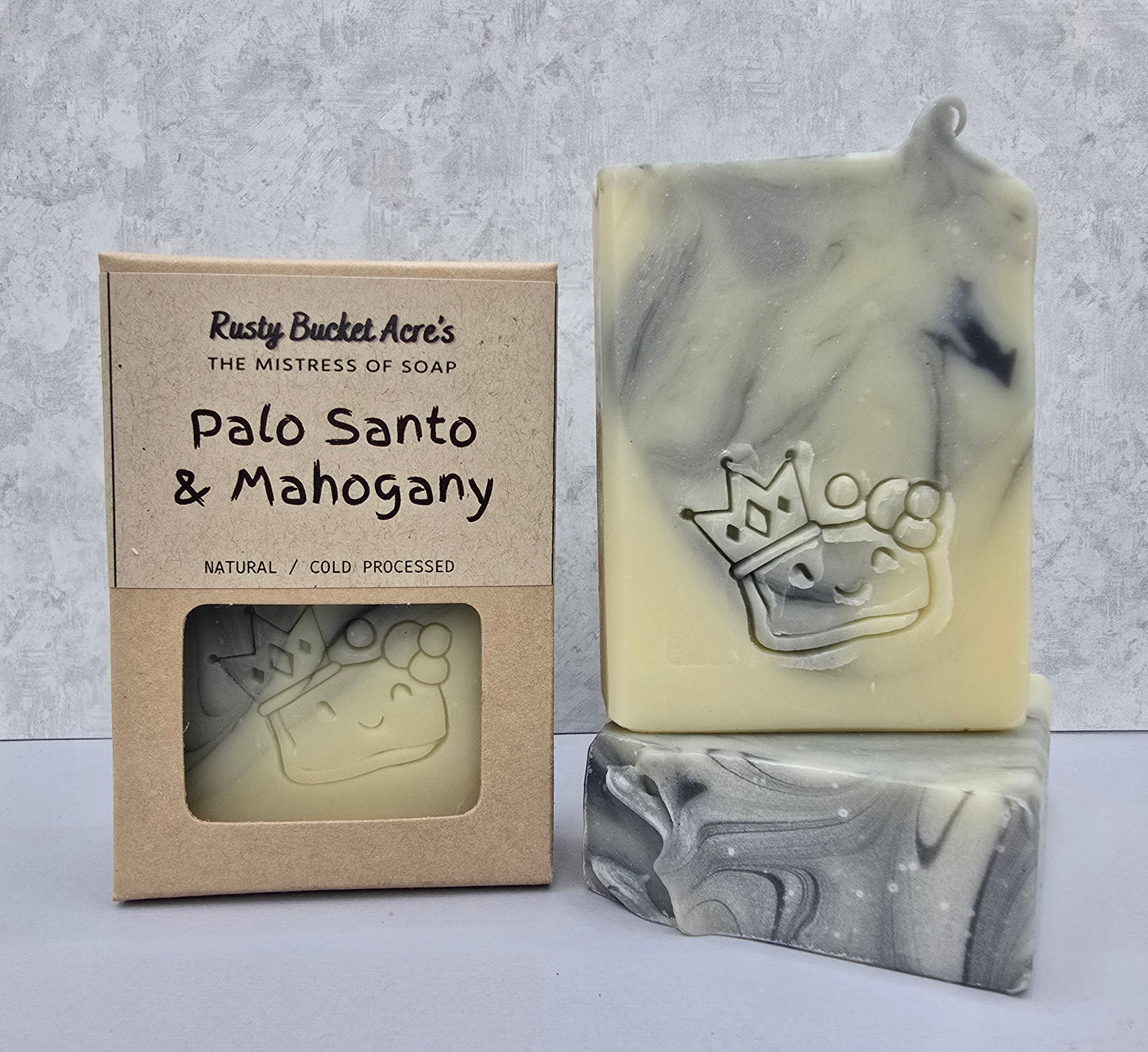 A handmade bar of soap in a cream and gray marbled pattern, displayed next to its kraft paper packaging box. The soap features an embossed crown and paw print design.