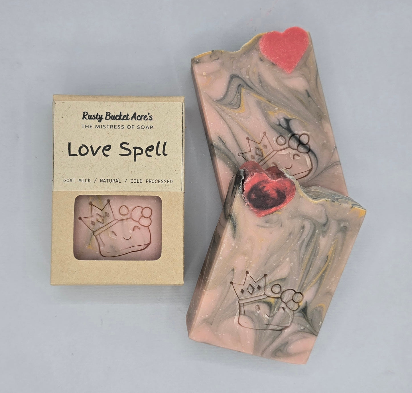 A handmade soap bar with pink, gray, and white marbled swirls, decorated with a crown and heart design, shown next to its kraft paper packaging box labeled 'Love Spell' by Risty Bucket Artie's.