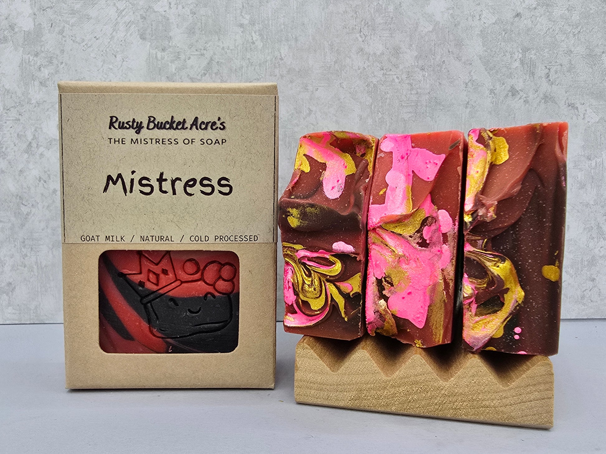 Three decorative handmade soap bars with pink, brown, and gold swirls displayed on a small stand next to their kraft paper packaging box labeled 'Rusty Bucket Acre's The Mistress of Soap Mistress'.